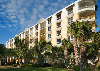 A modern multi-story residential building with yellow and white exterior, balconies, and large windows, surrounded by lush palm trees and green lawn under a partly cloudy blue sky.