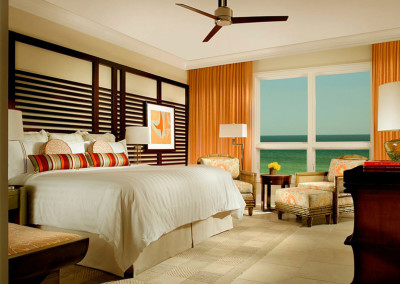 A luxurious bedroom features a large bed with a white duvet and colorful striped pillows. The room has a modern ceiling fan, cushioned chairs with matching patterns, a sea view through a large window, and warm-toned curtains. A framed artwork adorns the wall.