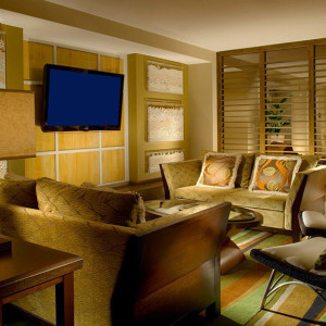 A cozy living room with a modern design featuring a large TV mounted on a yellow-paneled wall, surrounded by artwork. The space includes a beige couch with patterned pillows, a matching armchair, and a stylish coffee table. A wooden divider separates the areas.