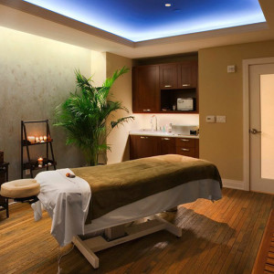 A serene massage room with a massage table covered in white and brown linens. The room has dim lighting, candles, a potted plant, wooden flooring, and cabinets with a microwave and sink. The atmosphere is calm and inviting, ideal for relaxation and therapy.