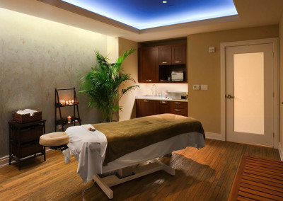 A serene massage room with a massage table covered in white and brown linens. The room has dim lighting, candles, a potted plant, wooden flooring, and cabinets with a microwave and sink. The atmosphere is calm and inviting, ideal for relaxation and therapy.